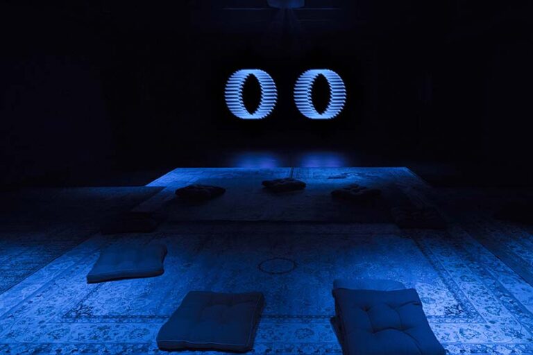 A dark room with two vibrant blue ellipses glowing on the wall. On the floor are pillows and oriental rugs.