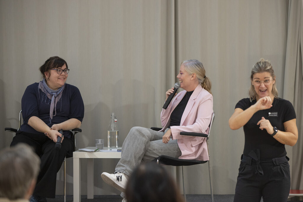 Två kvinnor sitter och samtalar på ett podie. En tredje kvinna syntolkar samtalet.
