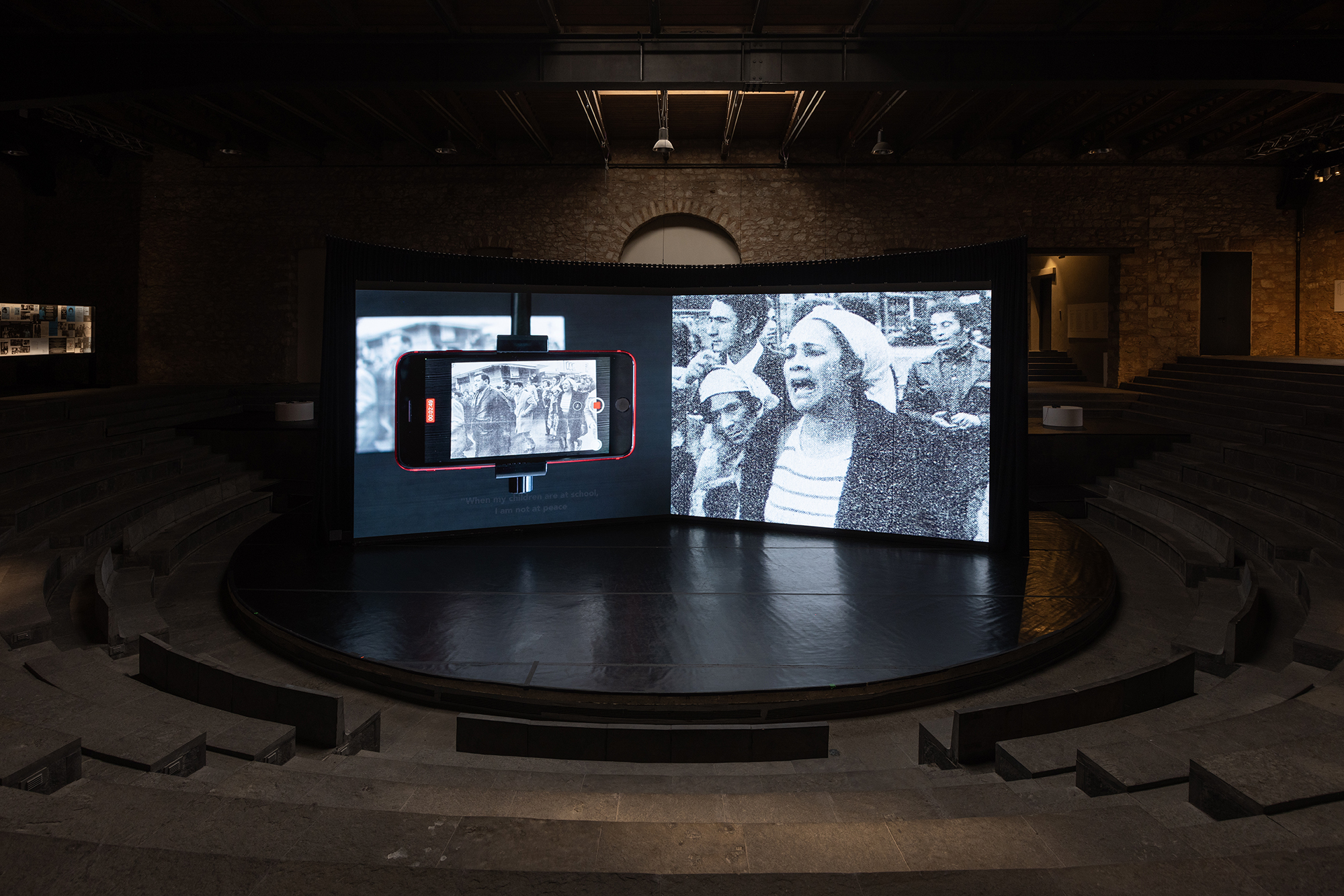 En dubbelsidig projektion på två skärmar i svartvitt av något som ser ut att vara en demonstration. Installationen visas på en scen i ett mörklagt rum.