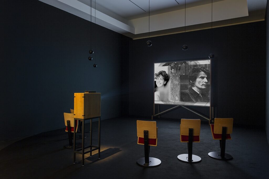 Ett mörklagt rum med tre träfärgade stolar framför en filmskärm med två svartvita porträtt. Bredvid stolen står en projektor i en låda.