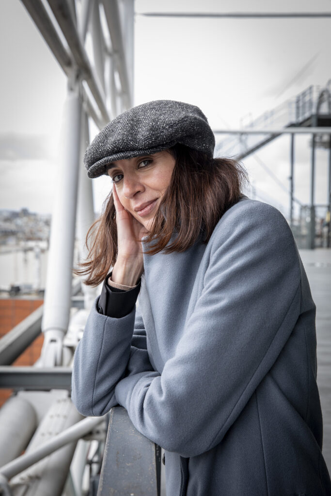 Konstnären Bouchra Khalili utomhus i grått höstväder iklädd en grå kappa och en grå basker, lutandes mot ett räcke.