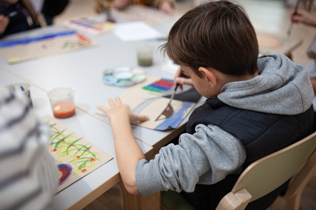 En pojke iklädd grå hoodie och väst sitter och målar med pensel. Man ser hans bakhuvud och hans teckning.