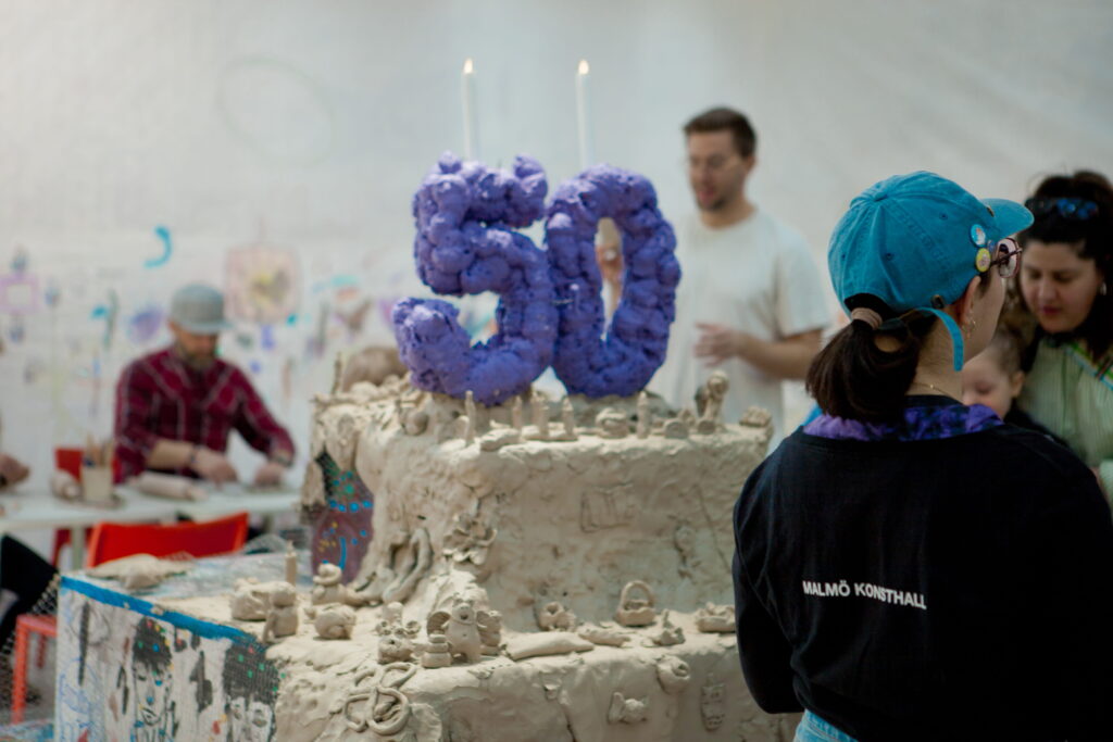 En tårta med siffran 50 skapad i lera och en person med Malmö Konsthall tröja i förgrunden.