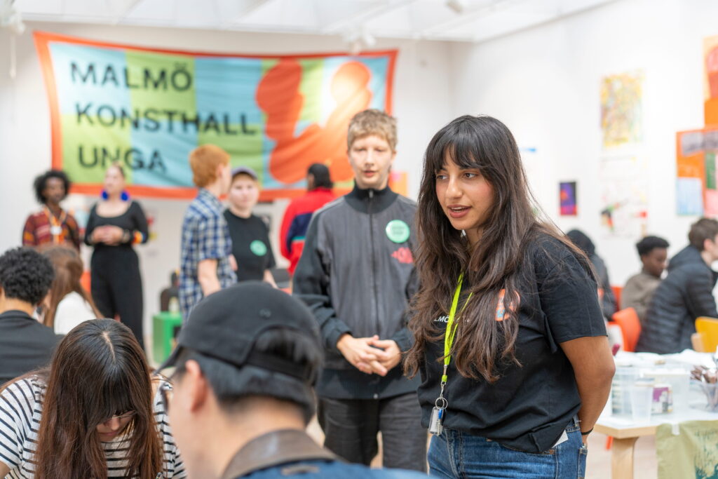 En person iklädd Malmö Konsthall kläder i Verkstan omgiven av ungdomar och en banderoll med texten 