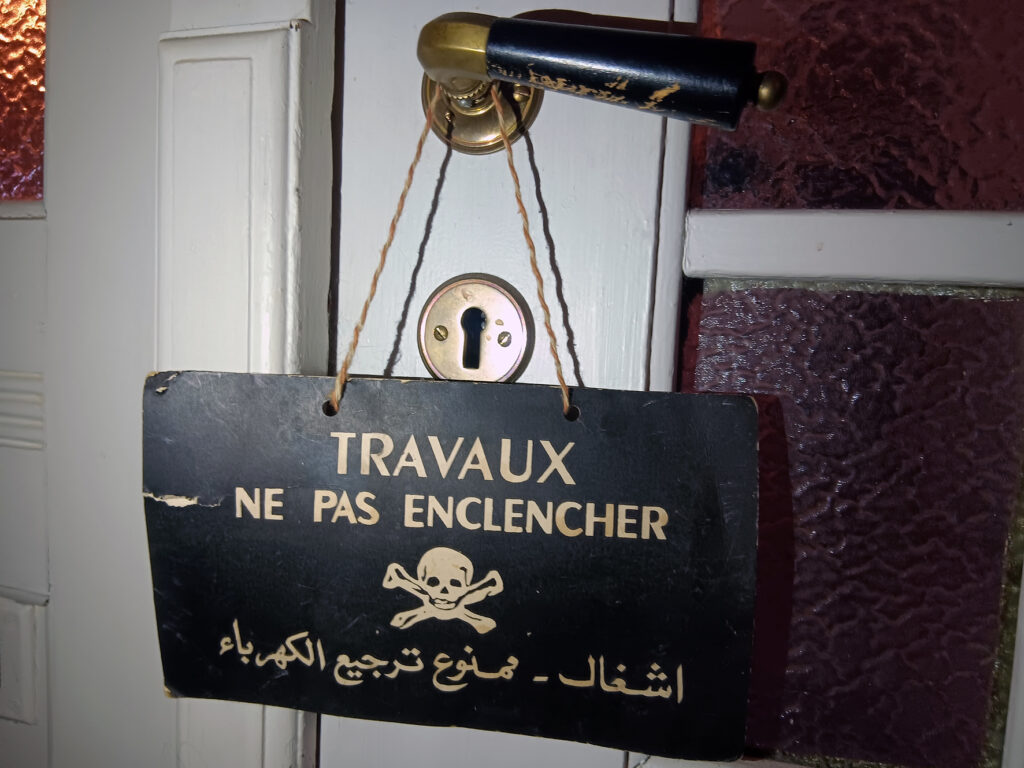 En svart skylt hänger på ett mässigfärgat dörrhandtag. Dörren är grå och har fönster med spröjs. Bilden är tagen med blixt.