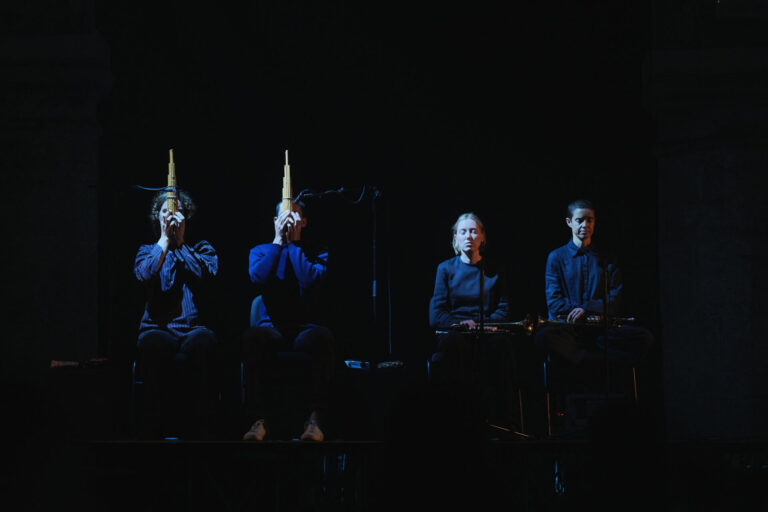 Fyra personer sitter på rad, mörkt klädda i ett mörkt rum. Kallt vitt ljus lyser upp deras ansikten.