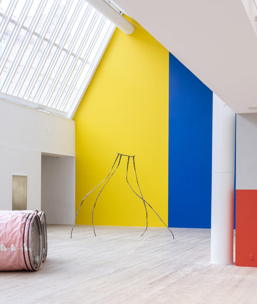 Installationsbild från utställningen Torrlägg Öresund, 2025. Ljusschaktets höga vägg är målad i IKEAs gul och blå nyanser. Framför väggen reser sig en av Hani Kamalis gängliga och långbenta metallskulpturer.