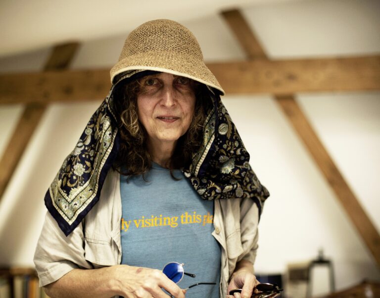 Porträtt av CC Hennix. Hon står iklätt blå t-shirt och vit skjorta i ett ljust rum. Hon har en stråhatt på huvudet med en scarf under som hänger över hennes axlar.