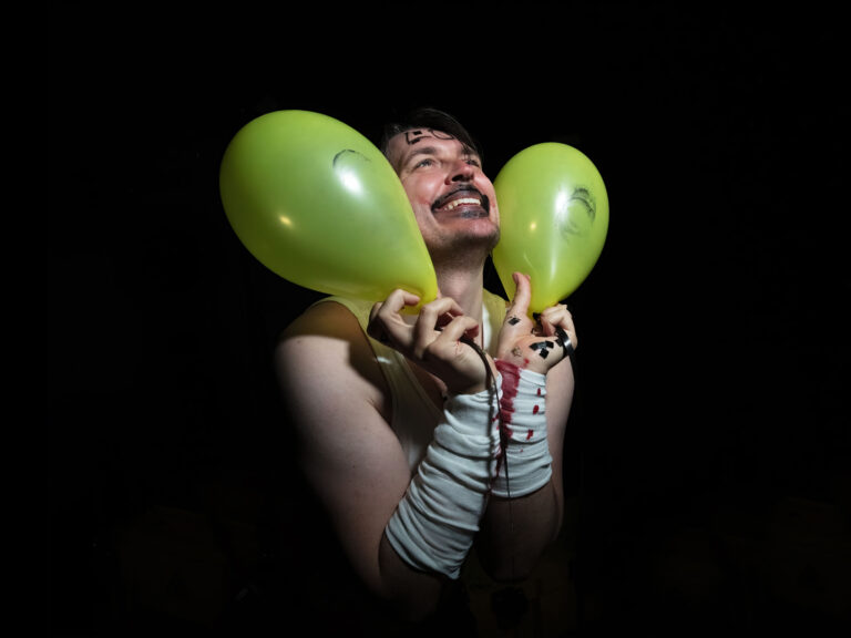 En person ler stort och blickar uppåt. Runt om ansiktet har han två gröna ballonger.