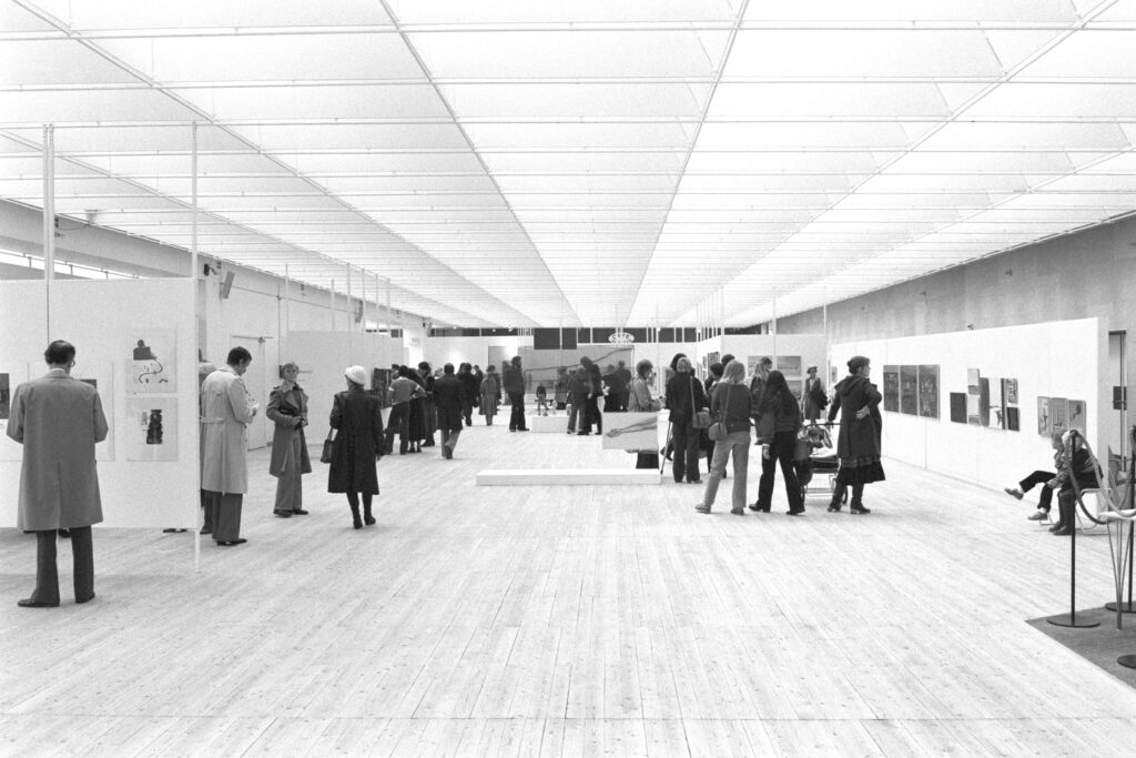 Utställningsvy under vernissage. Svartvit bild av personer som står och kollar på verken i hallen.