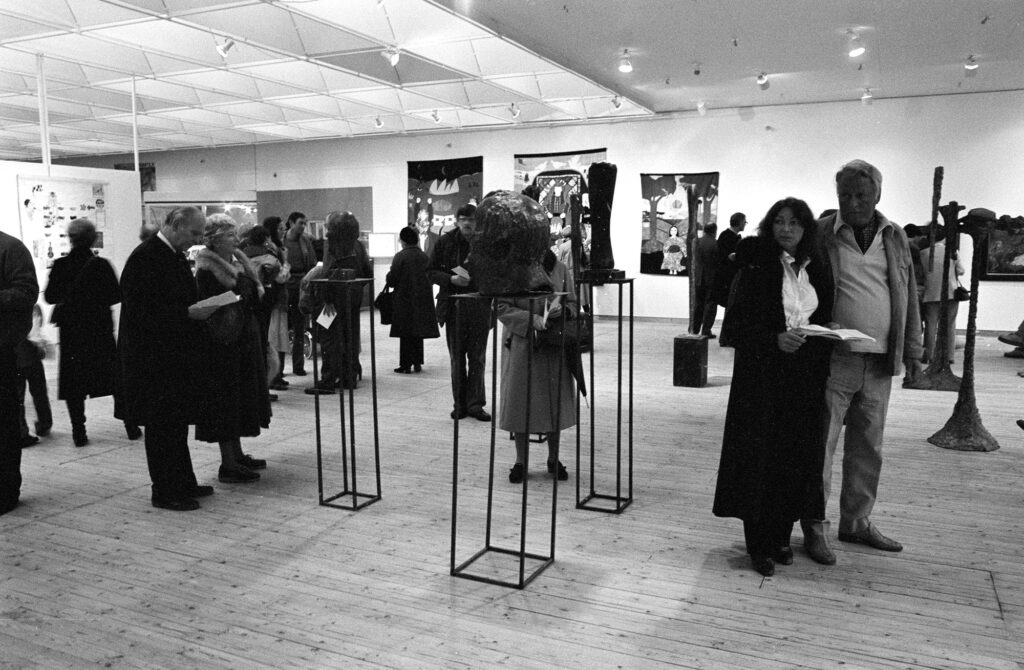 Svartvit bild från vernissagen av Skånes konstförenings Höstsalong 1980. Flera personer står i utställningshallen och kollar på verken.