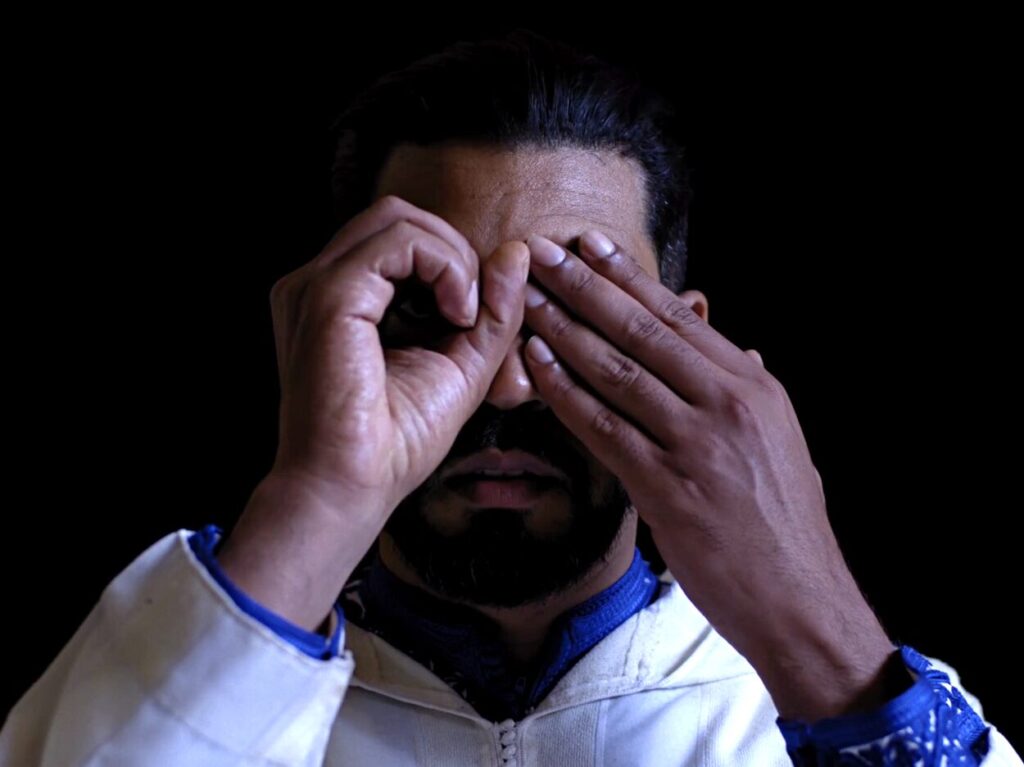 Stillbild från videoverk av konstnären Bouchra Khalili. En man formar handen som ett kikarsikte att titta igenom och håller för sitt andra öga.