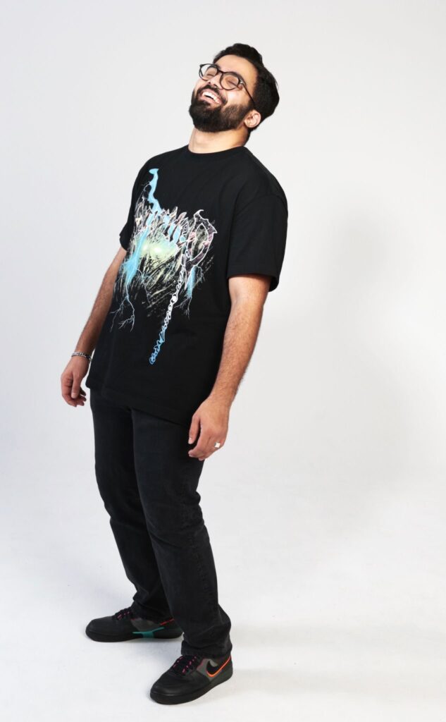En man står lätt bakåtlutad och skrattar stort. Han har mörkt hår, glasögon och en svar t-shirt med tryck.