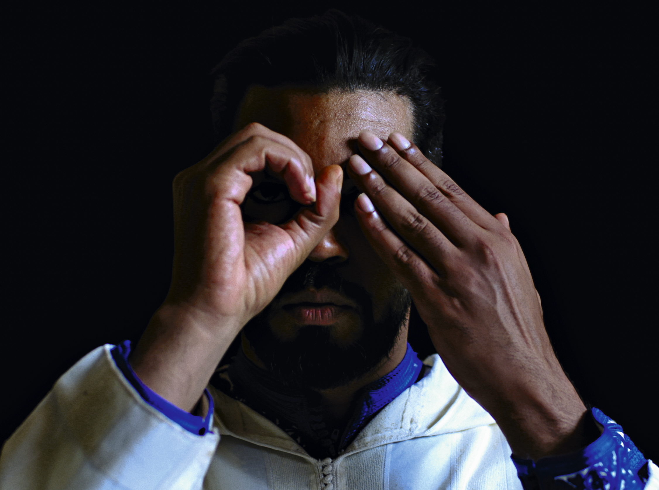 En man står i ett mörkt rum med en vit tröja. Han håller ena handen i formen av en cirkel framför ena ögat, och den andra handen hållandes för andra ögat.