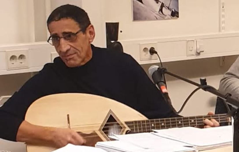 En man sitter vid ett bord med ett instrument i famnen. Han har mörkt hår och glasögon.
