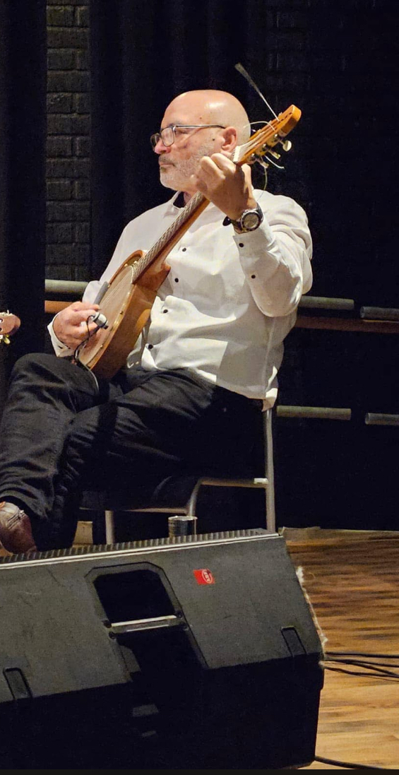 En man iklädd vit skjorta. Han sitter på en scen med en banjo i famnen och spelar.
