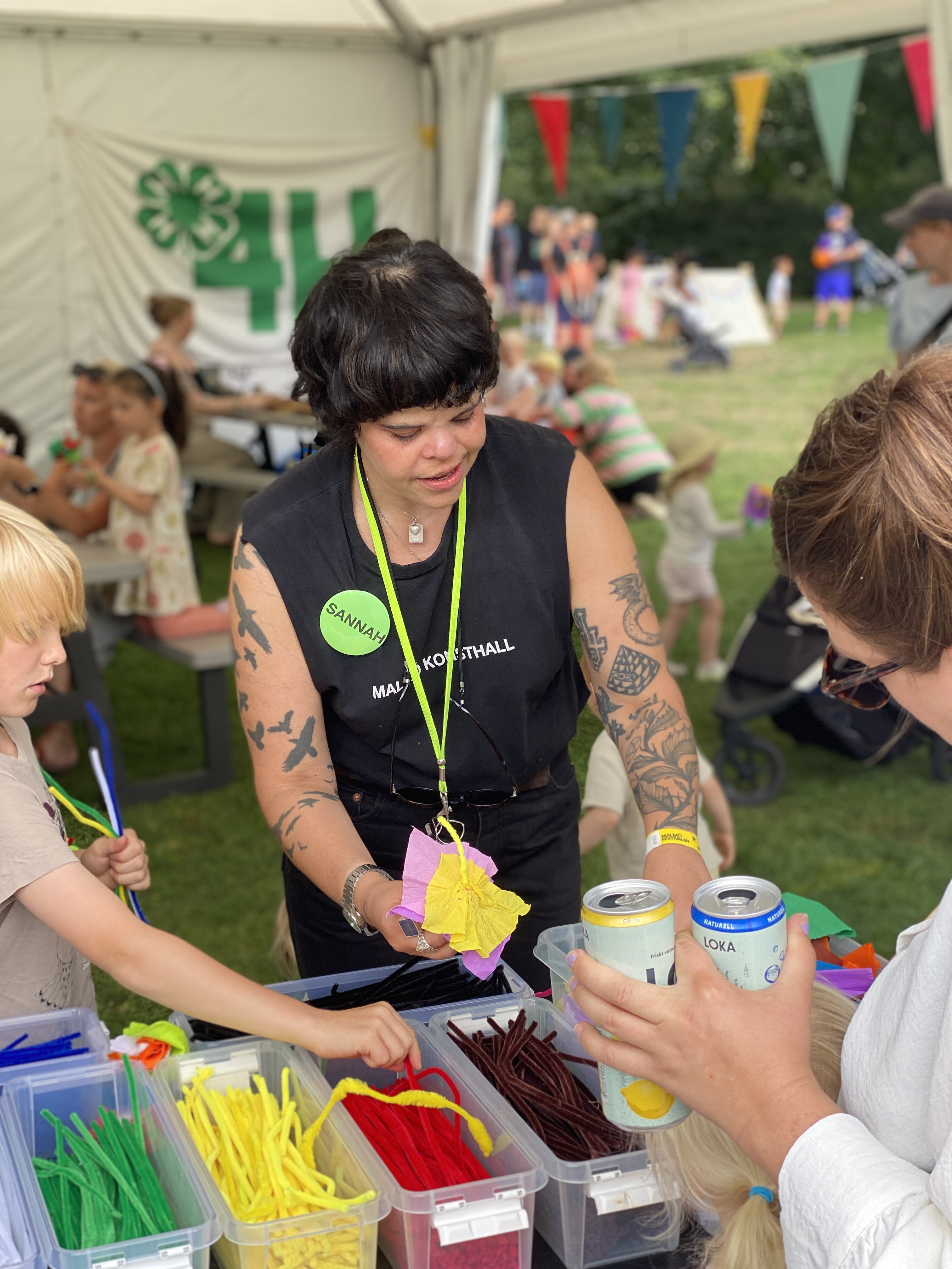 En konstpedagog står vid ett bord med piprensare och pysselmaterial i ett tält på Malmöfestivalen, några barn omkring henne.